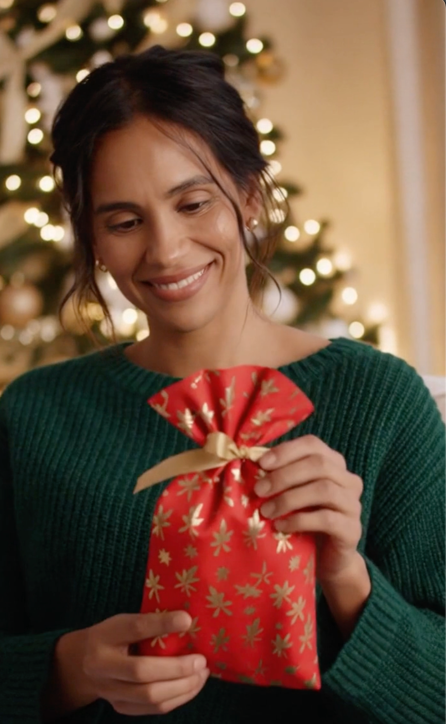 Woman holding a red gift bag with gold patterns in front of a Christmas tree.
