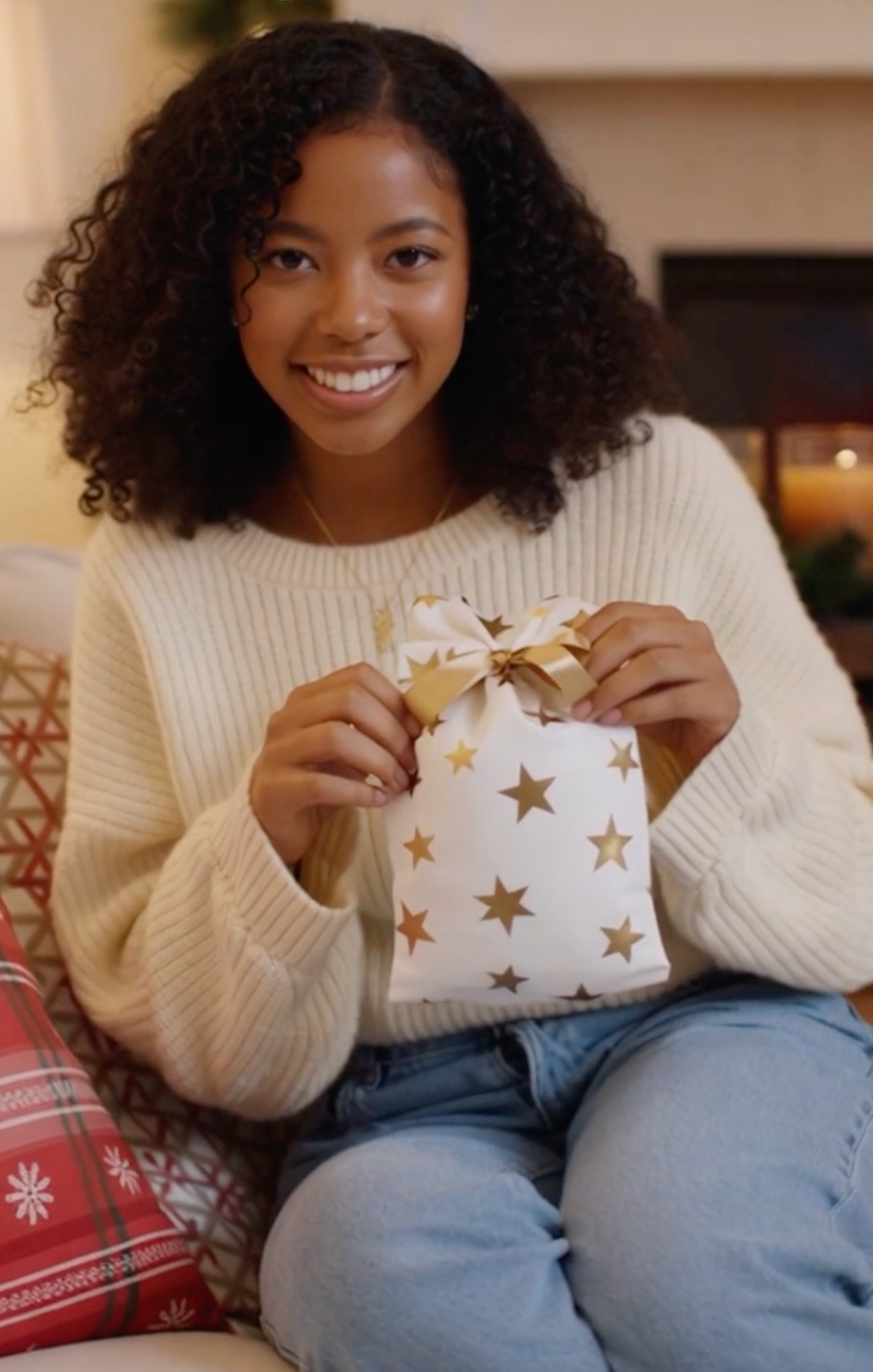 Woman holding a gift wrapped in paper with gold stars, sitting on a couch.